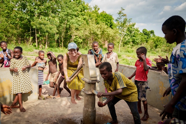 Eine Gruppe von Kindern steht mit einer erwachsenen Person an einer handbetriebenen Wasserpumpe im Freien. Die erwachsene Person betätigt die Pumpe, während ein Kind Wasser in einen Behälter füllt. Die anderen Kinder schauen lächelnd zu. Im Hintergrund stehen grüne Bäume, die Szene wirkt lebendig und gemeinschaftlich.